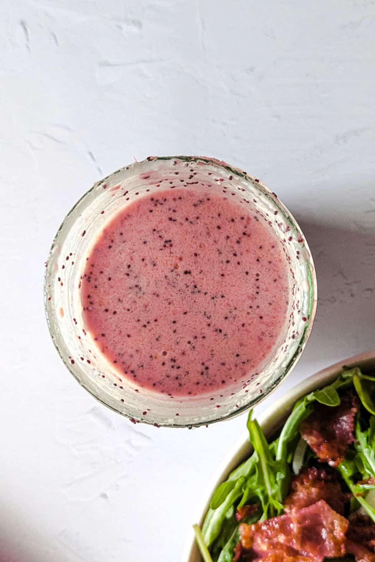 Top-down picture of small bowl of strawberry and poppy seed dressing alongside some spinach and arugula salad.