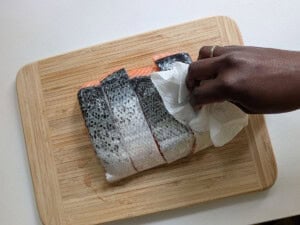 Gently patting a raw salmon fish dry with a paper towel on a cutting board.