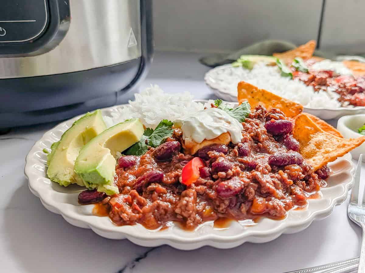 Plate of chili con carne topped with sour cream and served with avocado slices, rice, and tortilla chips.