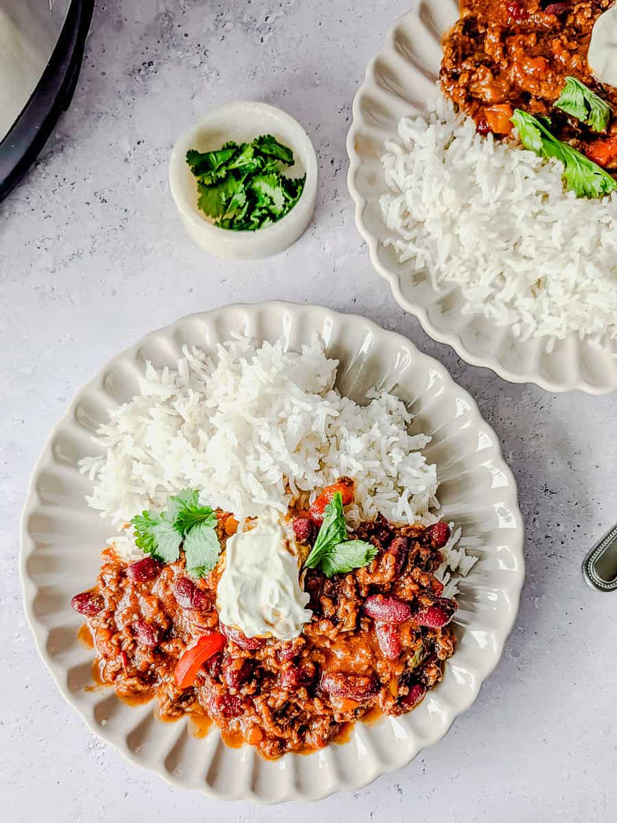 A top down image of chili con carne served with white rice and sour cream, garnished with fresh coriander.