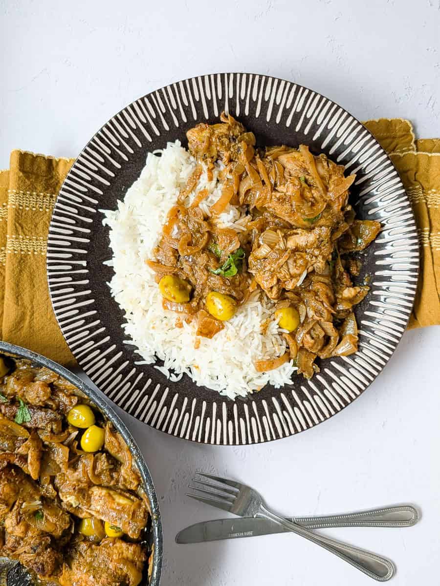 Chicken yassa served with white rice, caramelized onions, and green olives on a patterned plate.