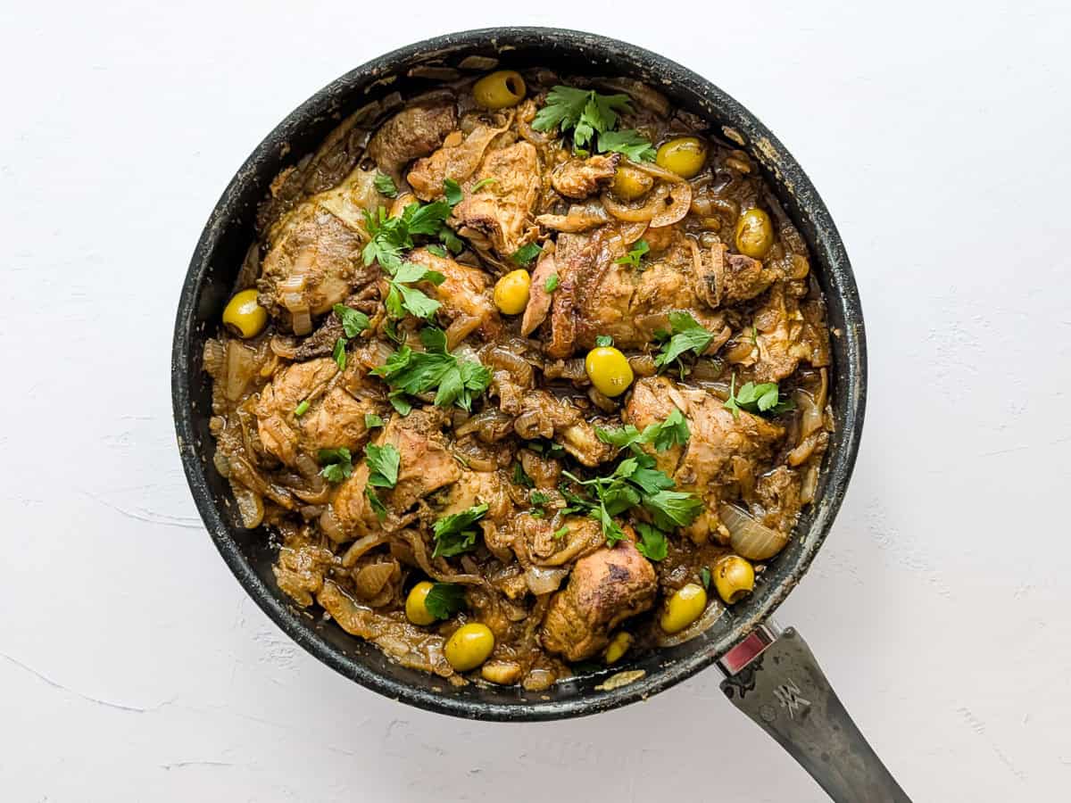 Top-down view of chicken yassa cooked in a pan with caramelized onions, olives, and parsley.