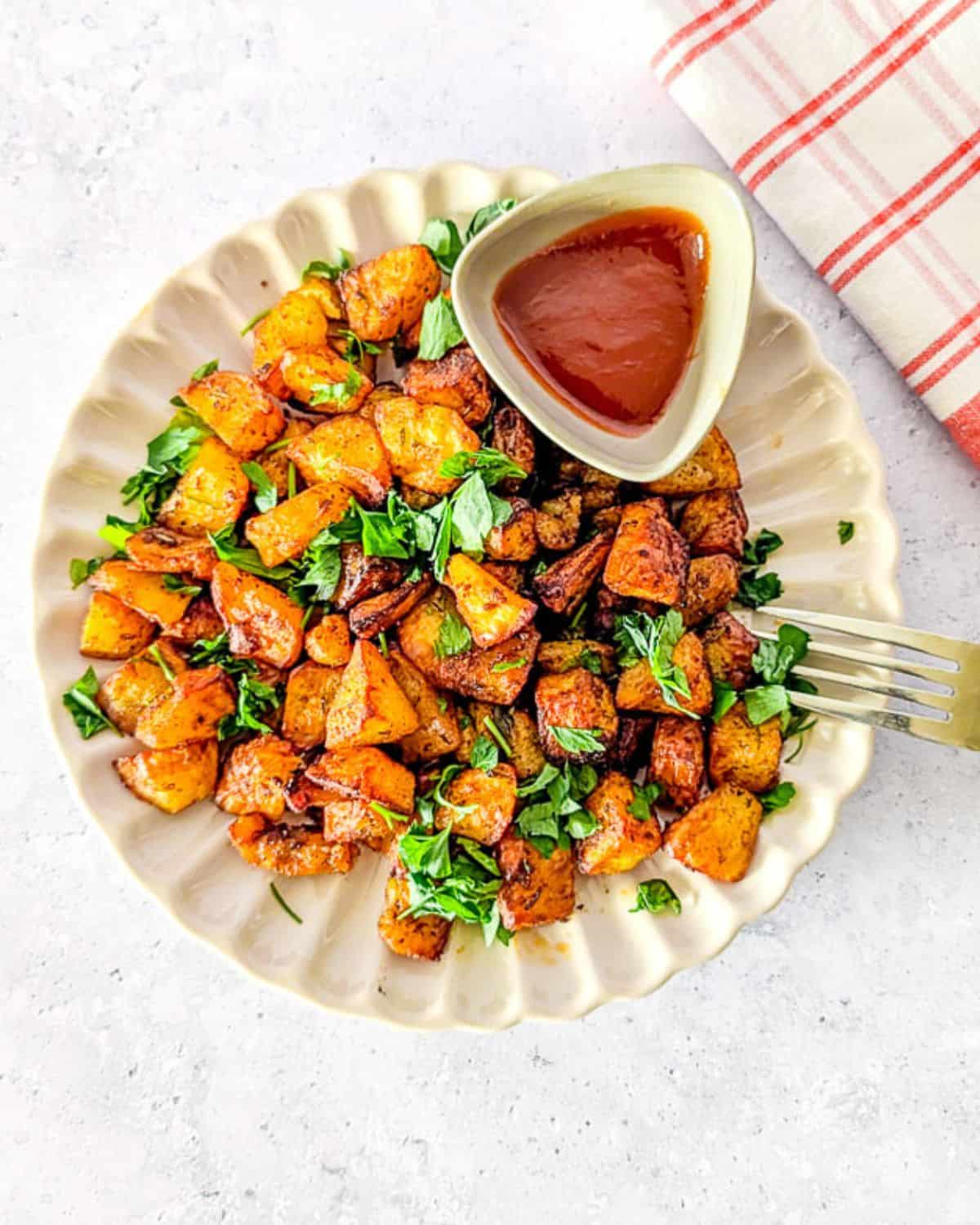 Overhead view of crispy diced air fryer potatoes topped with fresh herbs, served on a white scalloped plate with a small bowl of ketchup and a fork.