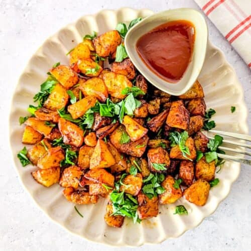 Overhead square shot of crispy air fryer diced potatoes garnished with herbs, plated with a small dish of ketchup and a fork resting on the side.