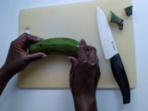 Hands holding a green plantain on a cutting board with the ends cut off.