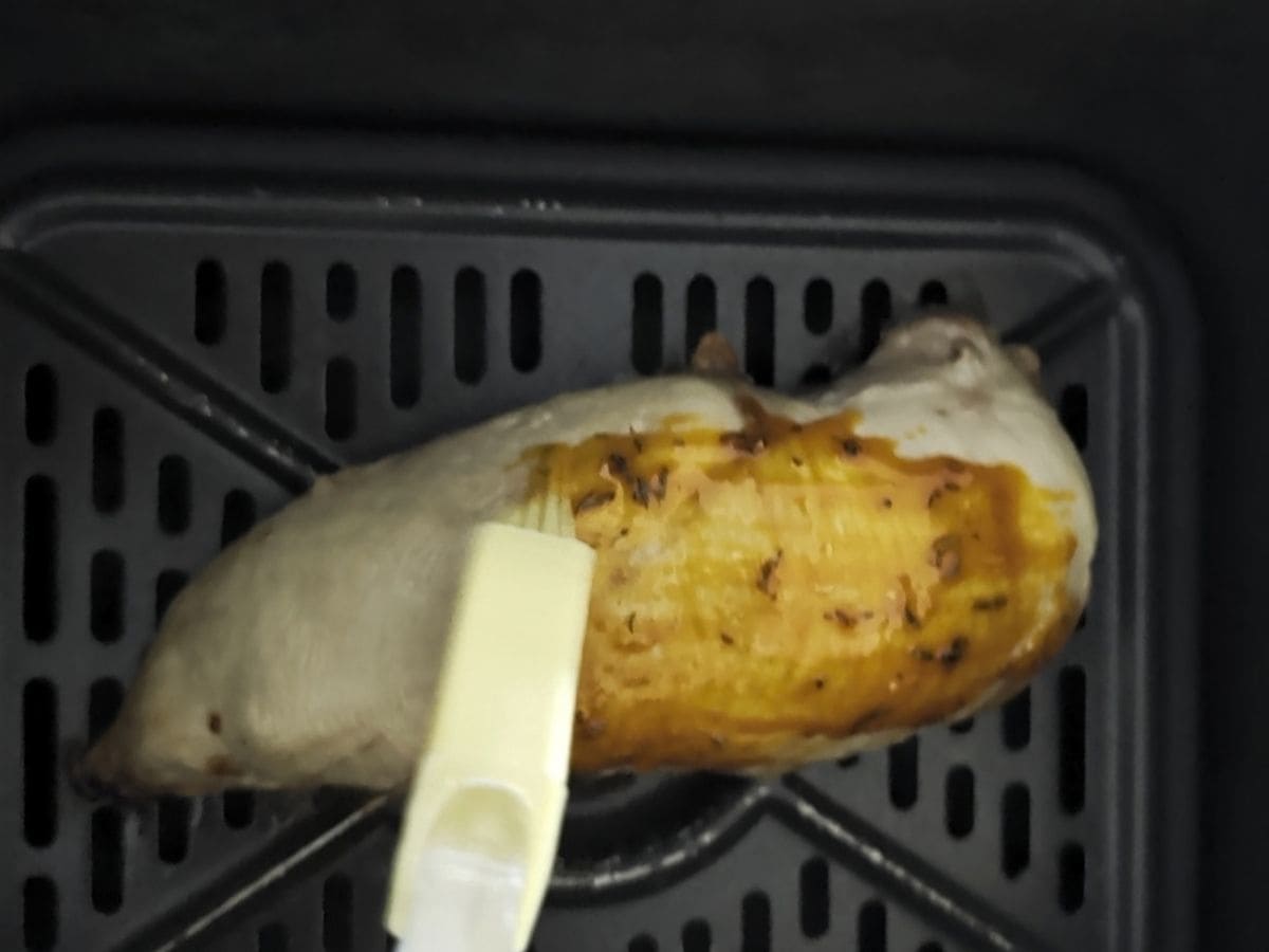 A close-up image of sliced chicken breast being brushed with a seasoning in an air fryer basket.