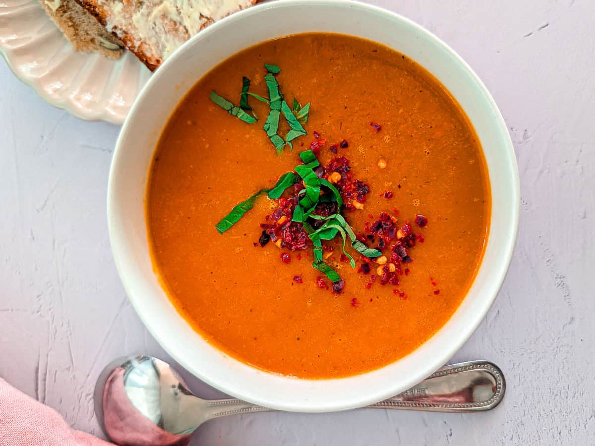 Overhead view of roasted tomato soup in a white bowl, garnished with fresh basil and chili flakes.