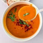 Bowl of roasted tomato soup garnished with fresh basil and chili flakes, with a spoonful of soup held above it.