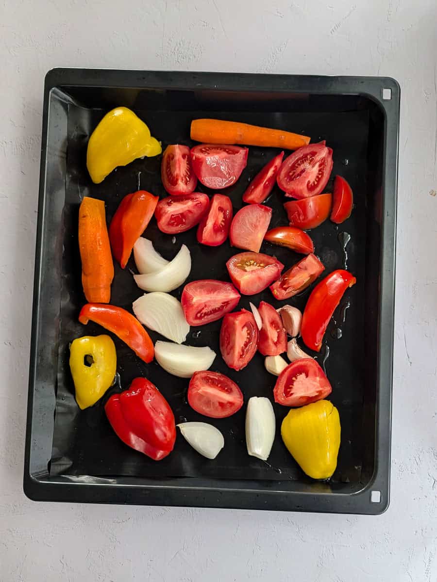 A top-down image of fresh tomatoes, bell peppers, carrots, onion, and garlic arranged on a baking tray.